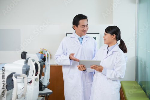 Cheerful Asian man and woman in white gowns of ophthalmology doctors sharing tablet in hospital cabinet