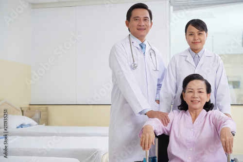 Cheerful Asian man and woman in medical gowns standing with mature woman in wheelchair in hospital ward smiling at camera