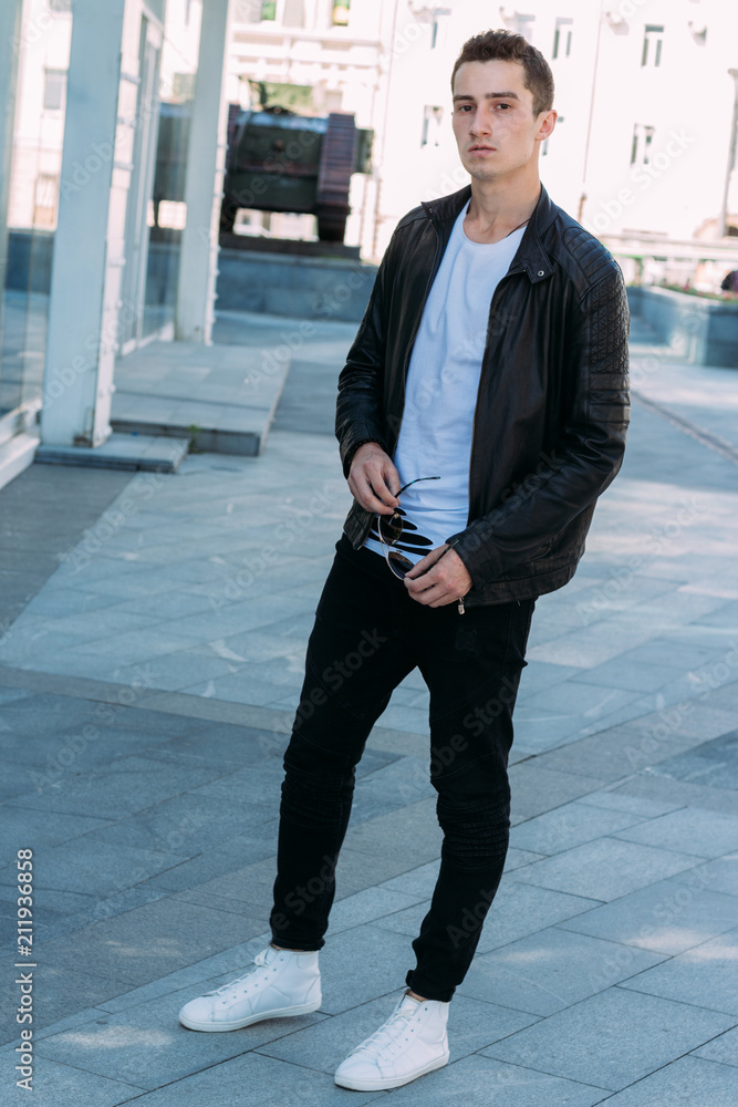 Stockfoto beautiful and attractive guy is standing in the city, posing.  dressed in a black leather kurta, white T-shirt, jeans and white shoes. |  Adobe Stock