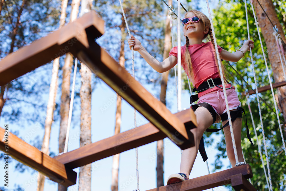 Fototapeta premium Exciting adventure. Pleasant little girl moving around rope park confidently and enjoying herself