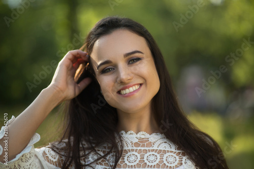 Portrait of sweet girl smiling. Stunning beautiful lady dressed in white laced dress smiles and fixes her hair behind her ear with her hand. Forest in background. © Svyatoslav Lypynskyy