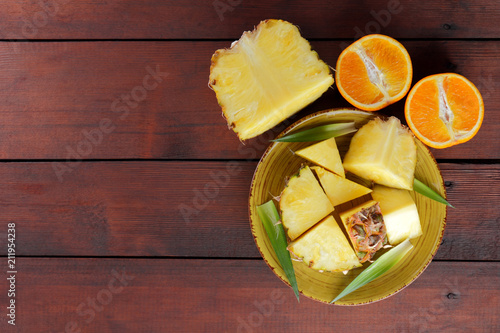 Pieces of pineapple and oranges on a yellow plate on wooden boards, pineapple and halves of orange on a wooden background, salad of tropical fruits for breakfast, vegetarian food, copy space