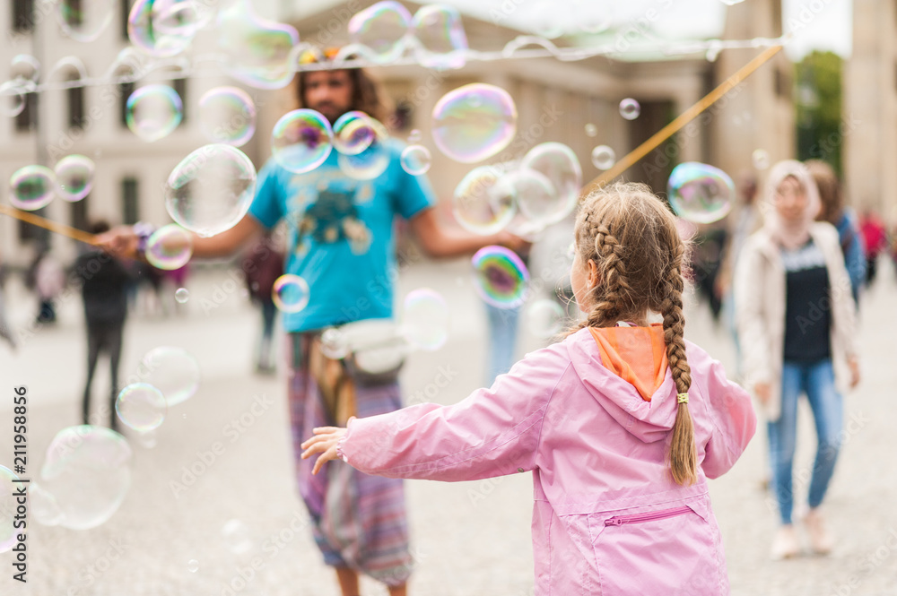 Fototapeta premium Street performer, busker, entertaining the crowd in front of the Brandenburg Gate in Berlin on an overcast summer day. Children playing with colorful soap bubbles floating in the foreground.