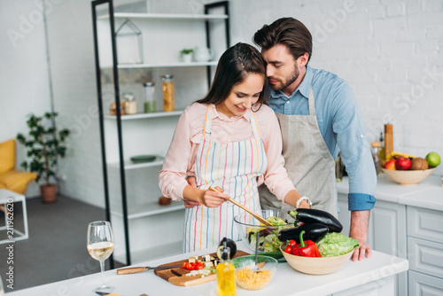 girlfriend cooking and mixing salad in kitchen and boyfriend hugging her