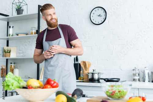 handsome smiling bearded man in apron looking away while cooking in kitchen