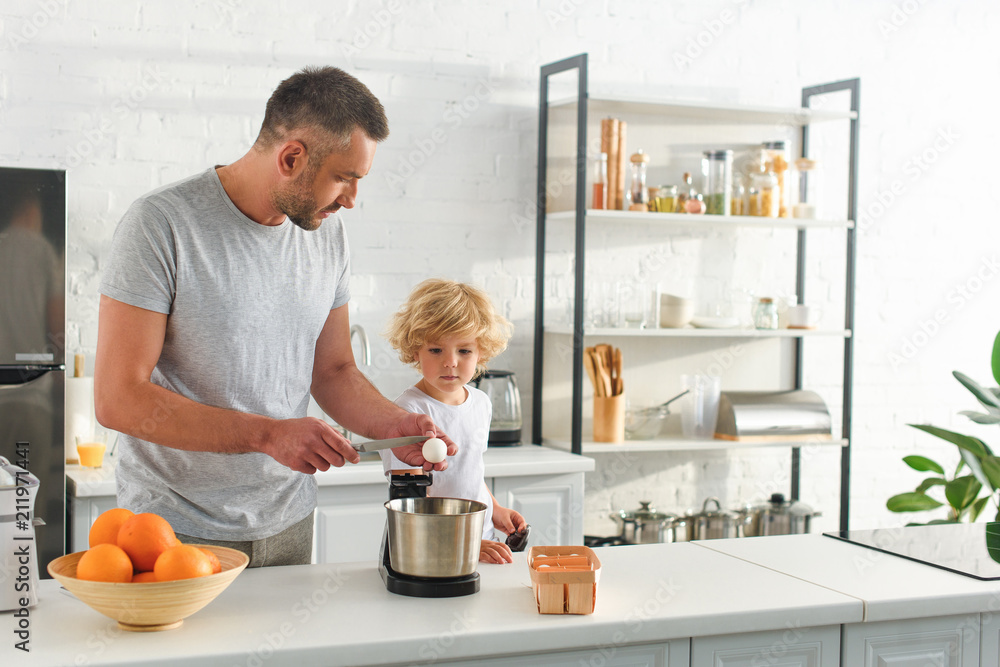 side view of man breaking egg by knife for making dough while his son standing near at kitchen side view of man breaking egg by knife for making dough while his son standing near at kitchen