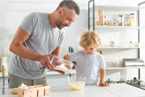 man pouring flour into bowl while his son whisking eggs at kitchen