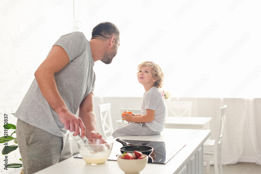 father whisking dough in bowl and talking to smiling son while he sitting on tabletop at kitchen father whisking dough in bowl and talking to smiling son while he sitting on tabletop at kitchen