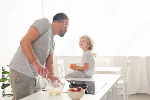 father whisking dough in bowl and talking to smiling son while he sitting on tabletop at kitchen