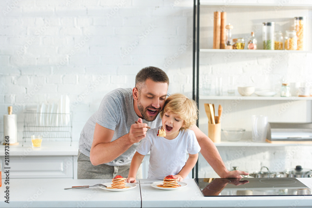 happy father and son eating pancakes at kitchen happy father and son eating pancakes at kitchen