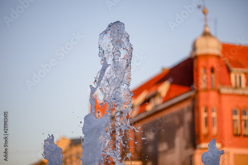 Fountain spurt, water drops, main street in the city as a background. Evening sun light on the facades - old town in Gniezno, Poland.  photo