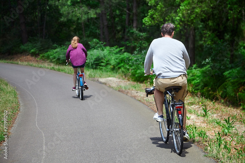 Fototapeta Naklejka Na Ścianę i Meble -  Loving married couple riding on bicycles. A man in beige shorts and a woman in a purple shirt. View from the back. Bike ride in the nature on a sunny summer day