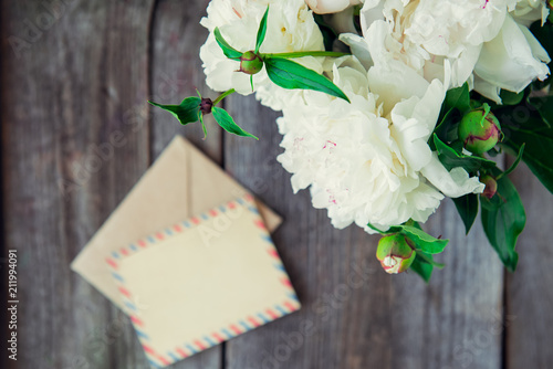 Focused White peony flowers bouquet and blurred blank greeting card and craft paper envelope on the old wooden rustic table background. Postcard mock up. Selective focus. Copy space.
