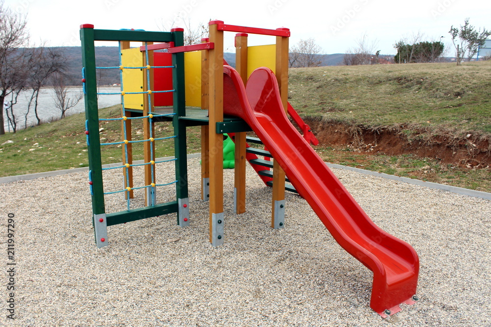 Outdoor public playground equipment with red slide, steps and climbing net surrounded with playground sand, sea and winter vegetation in background Outdoor public playground equipment with red slide, steps and climbing net surrounded with playground sand, sea and winter vegetation in background