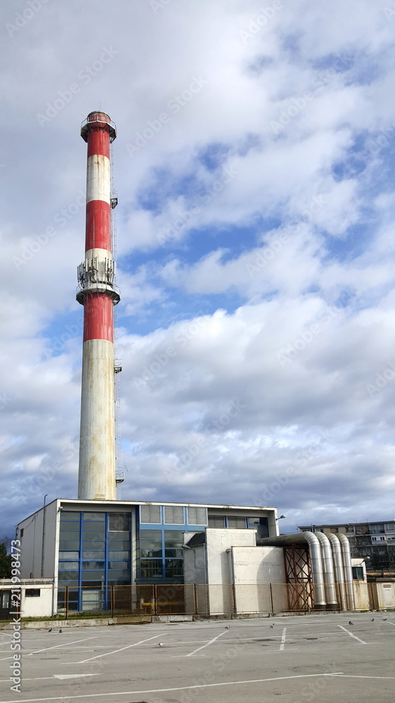 Old small power plant building with high industrial chimney and steel pipes on a cloudy day with completely empty parking in front Old small power plant building with high industrial chimney and steel pipes on a cloudy day with completely empty parking in front