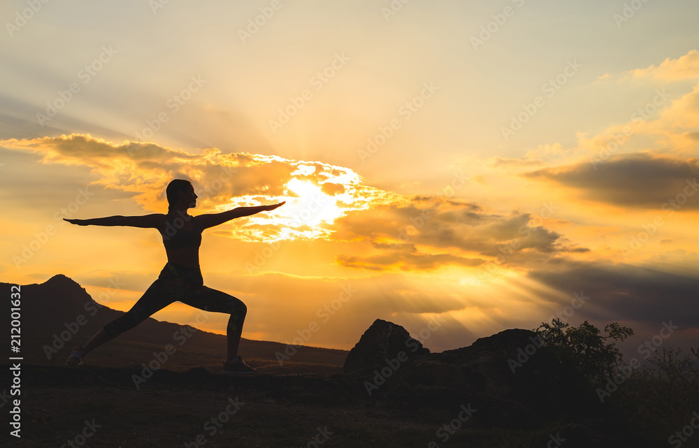 Silhouette of young woman practicing yoga or pilates at sunset or sunrise in beautiful mountain location, doing lunge exercise, standing in Warrior.