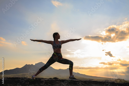 Silhouette of young woman practicing yoga or pilates at sunset or sunrise in beautiful mountain location, doing lunge exercise, standing in Warrior.