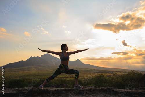 Silhouette of young woman practicing yoga or pilates at sunset or sunrise in beautiful mountain location, doing lunge exercise, standing in Warrior.