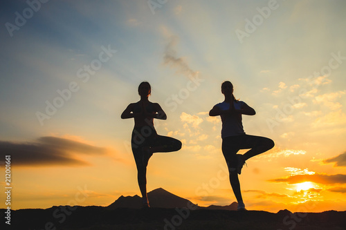 Silhouette of young womans practicing yoga or pilates at sunset or sunrise in beautiful mountain location.
