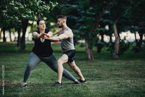 Fototapeta Naklejka Na Ścianę i Meble -  Morning training, sport, weight loss, teamwork and healthy lifestyle concept. Overweight woman doing yoga exercise with personal trainer support. Side or lateral lunge fat burning exercise
