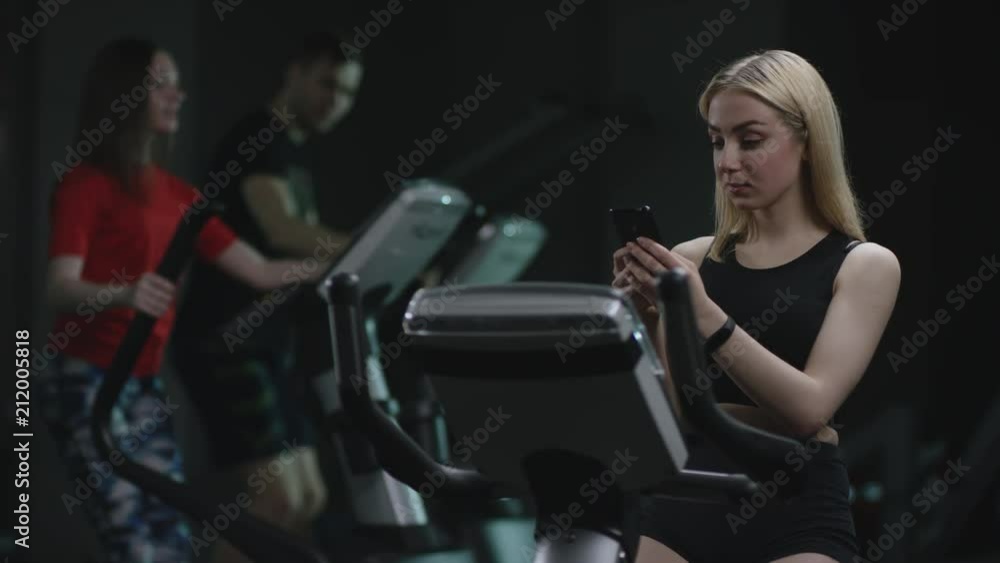 cute blonde girl is diligent working on an exercise bike and making selfie on her phone on a background of practiced girls cute blonde girl is diligent working on an exercise bike and making selfie on her phone on a background of practiced girls
