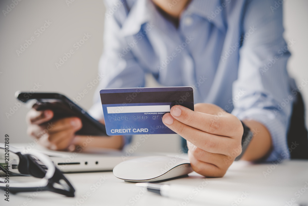 Fototapeta premium Hand holding a credit card in their hands and find information about a product using their mobile device to make purchases online and conduct financial transactions.