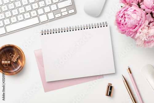 feminine workspace / desk with blank open notepad, keyboard, stylish office / writing supplies and pink peonies on a white background, top view photo