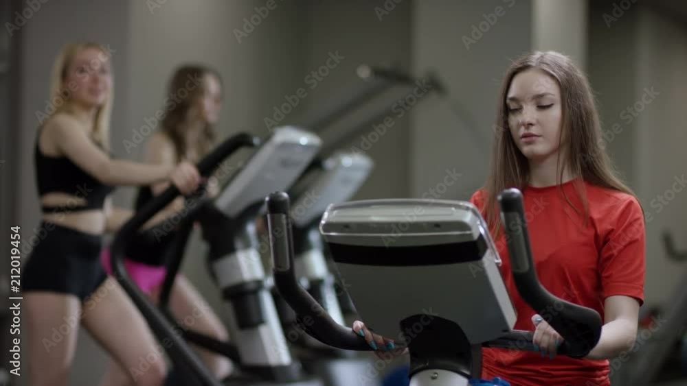 awesome girl in red shirt vigorously work on exercise bike and take a bottle of water to drink in the new gym against the background of engaged girls awesome girl in red shirt vigorously work on exercise bike and take a bottle of water to drink in the new gym against the background of engaged girls