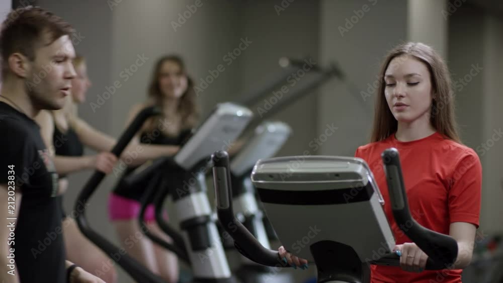 nice girl in red shirt vigorously work on exercise bike and trainer come greed her in the new gym against the background of engaged girls nice girl in red shirt vigorously work on exercise bike and trainer come greed her in the new gym against the background of engaged girls