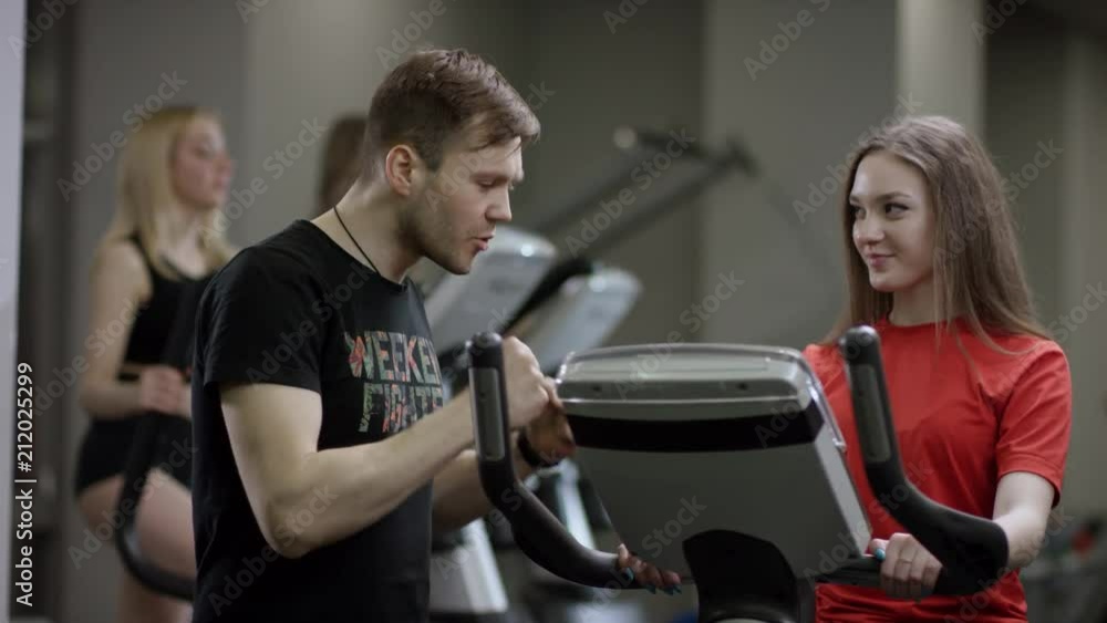 nice girl in red shirt vigorously work on exercise bike and trainer come help with motivation her in the new gym against the background of engaged girls nice girl in red shirt vigorously work on exercise bike and trainer come help with motivation her in the new gym against the background of engaged girls