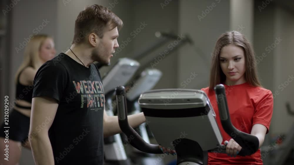 beautiful girl in red shirt vigorously work on exercise bike and trainer come help with motivation her in the new gym against the background of engaged girls beautiful girl in red shirt vigorously work on exercise bike and trainer come help with motivation her in the new gym against the background of engaged girls