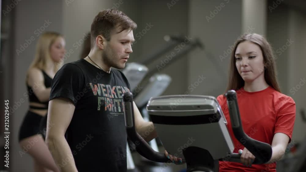 awesome girl in red shirt vigorously work on exercise bike and coach come instructs on the correct execution of the exercise in the new gym against the background of engaged girls awesome girl in red shirt vigorously work on exercise bike and coach come instructs on the correct execution of the exercise in the new gym against the background of engaged girls