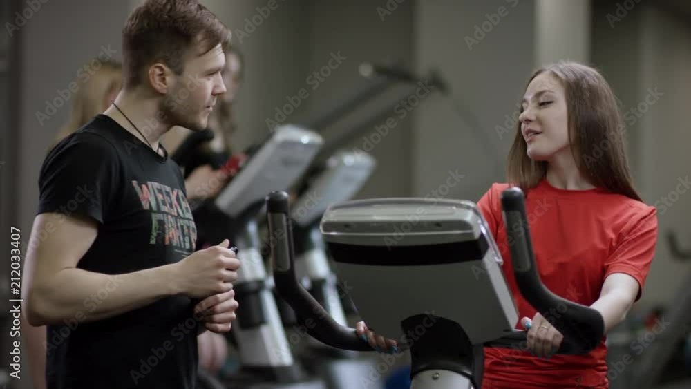 awesome girl in red shirt vigorously works on exercise bike and guy comes to take her number in the new gym against the background of engaged girls awesome girl in red shirt vigorously works on exercise bike and guy comes to take her number in the new gym against the background of engaged girls