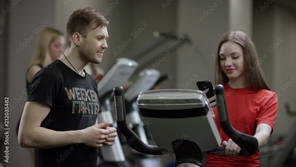 lovely girl in red shirt vigorously works on exercise bike and guy comes to take her number in the new gym against the background of engaged girls lovely girl in red shirt vigorously works on exercise bike and guy comes to take her number in the new gym against the background of engaged girls