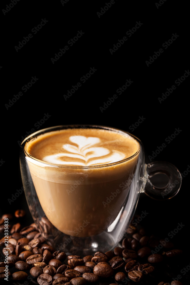 Cappuccino with a beautiful pattern of milk on top in a cup of double-sided glass on a black background, beside scattered coffee beans. Background image. Copy space, selective focus Cappuccino with a beautiful pattern of milk on top in a cup of double-sided glass on a black background, beside scattered coffee beans. Background image. Copy space, selective focus