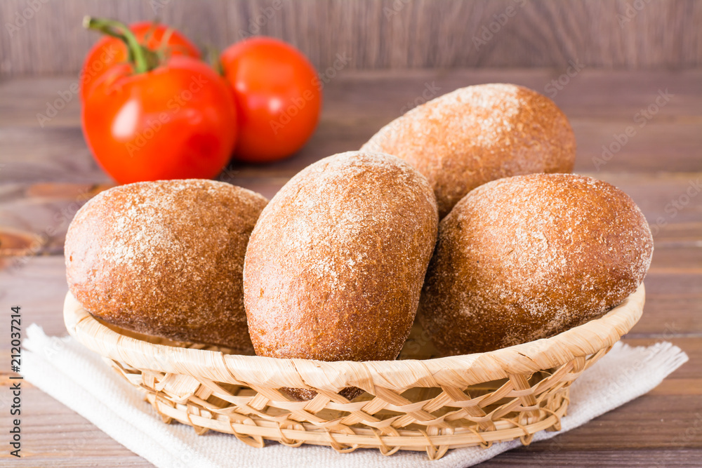 Fresh rye buns in a basket and tomato on a wooden table