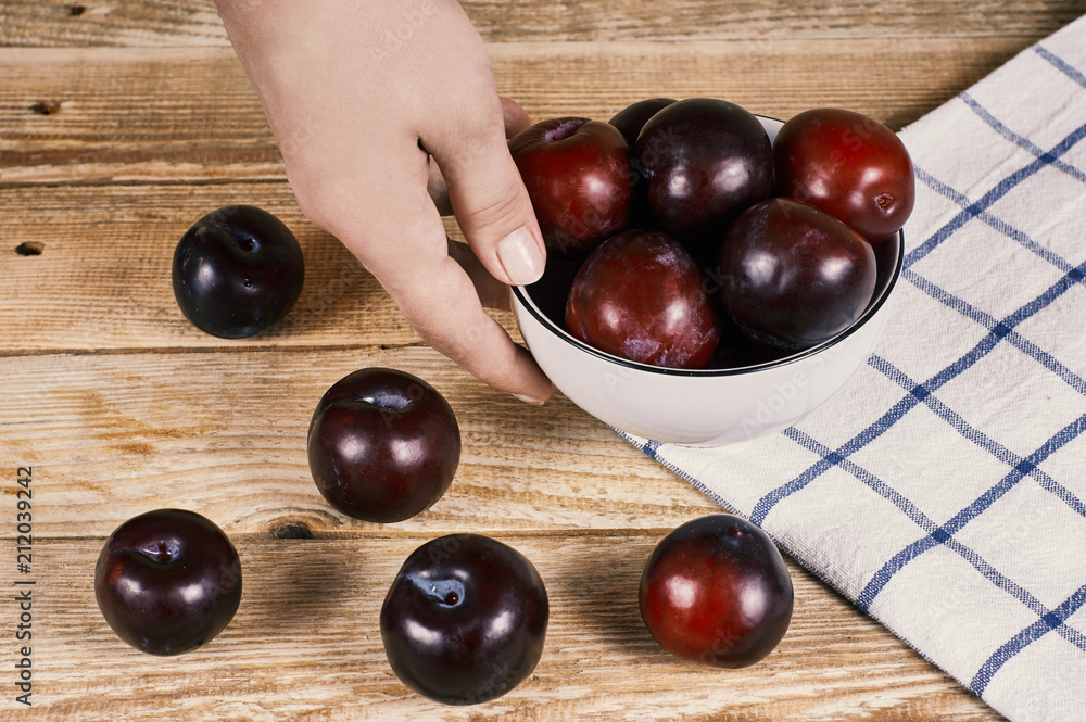 female hand holds a bowl with red plums next to a doily and a few plums in front, on a wooden background female hand holds a bowl with red plums next to a doily and a few plums in front, on a wooden background