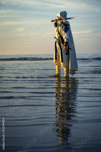 Portrait of assassin in white costume with the sword at the sea. He is posing...
