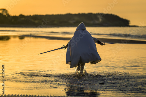 Portrait of assassin in white costume with the sword at the sea. He is posing...