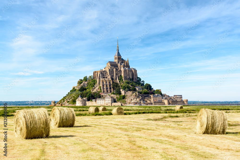 Obraz premium View of the Mont Saint-Michel tidal island, situated in France on the border between Normandy and Brittany, with round bales of straw in a field in the foreground under a blue sky with fibrous clouds.