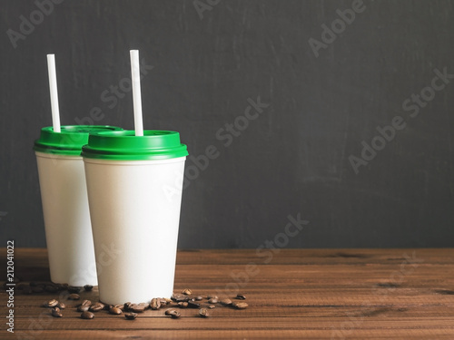 two white plastic coffee cups with a green lid on a wooden table with scattered coffee beans, copy space