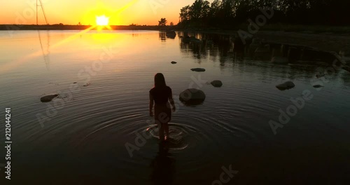Girl at the sea, C4K aerial view over a woman enjoying the sunset on a beach and cooling her feet in the water of the sea, on a sunny summer evening dusk, Hanko, Uusimaa, Finland