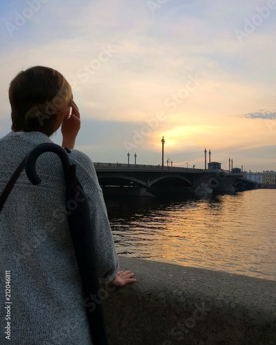 A girl with short hair stares into the distance to the sunset, next to the bridge across a large river. Background image. Silhouette on sunset background.