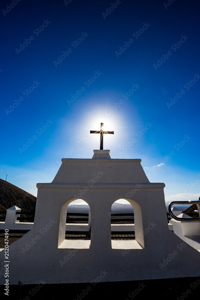 Hot Sun coming through the famous black Christian cross at the beautiful spot called Mirador De Femes in Lanzarote, Canary Islands, Spain. Two white windows and clear blue spring sky background Hot Sun coming through the famous black Christian cross at the beautiful spot called Mirador De Femes in Lanzarote, Canary Islands, Spain. Two white windows and clear blue spring sky background