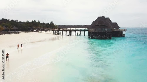 Flying over African traditional wooden house with pier bridge on stilts with thatched roof. Wide paradise beach with white sand and azure ocean on cloudy day. Nungwi and Kendwa beach, Zanzibar, Tanzan photo
