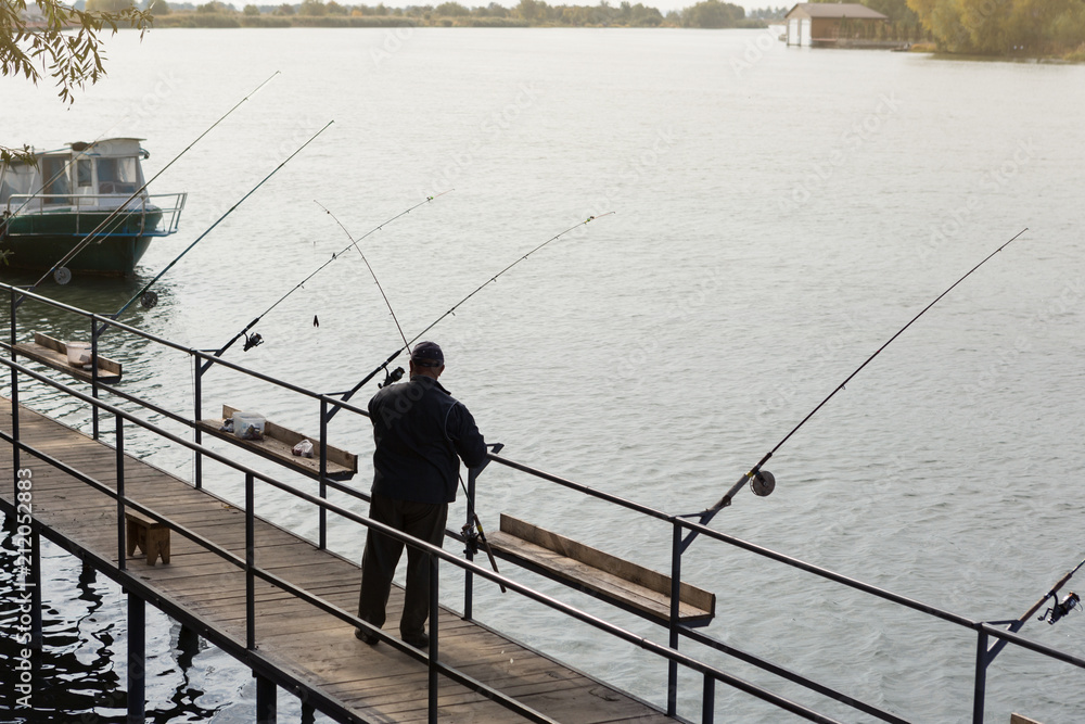 a fisherman on a wooden bridge catches fish in the river, many fishing rods stand in a row, early morning