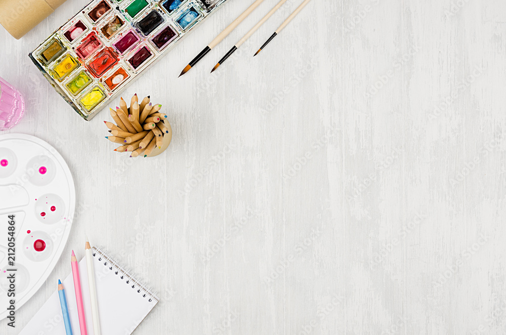 Fototapeta premium Workplace for drawings - watercolor paints, palette, brushes, colored pencils on white wood board, top view, copy space.