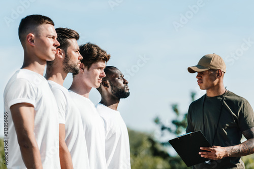 side view of african american tactical instructor with notepad and young soldiers on range