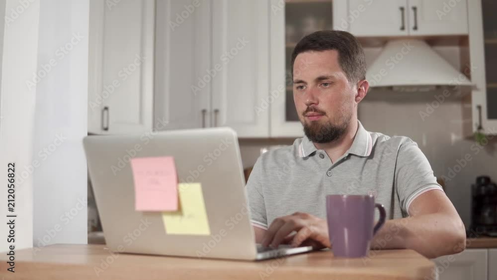 Adult bearded man sitting at the table with a laptop in kitchen and drinking tea. Man working on laptop indoor. Notes on laptop. Adult bearded man sitting at the table with a laptop in kitchen and drinking tea. Man working on laptop indoor. Notes on laptop.