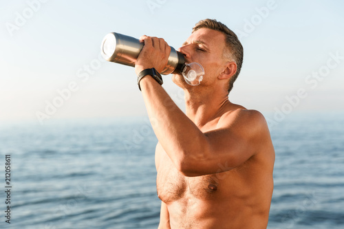 Handsome strong mature sportsman drinking on the beach.